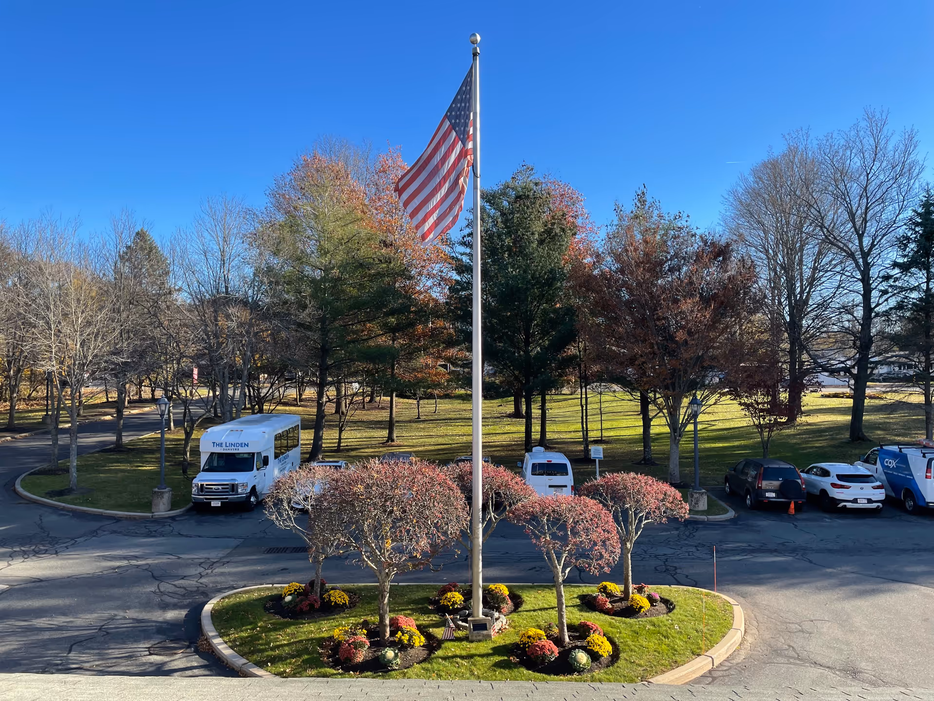 A circular landscaped area with small trees and flowers surrounding a tall flagpole flying the American flag. Behind the circle is a parking lot with several vehicles, including a white shuttle bus labeled 'THE LINDEN'. Trees with autumn foliage and a clear blue sky are visible in the background.