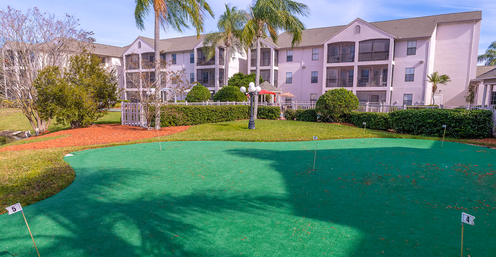 Outdoor view of Regency Residence - Sky Active Living showing a putting green with numbered flags, surrounded by green grass, palm trees, bushes, and a three-story residential building in the background under a clear sky.