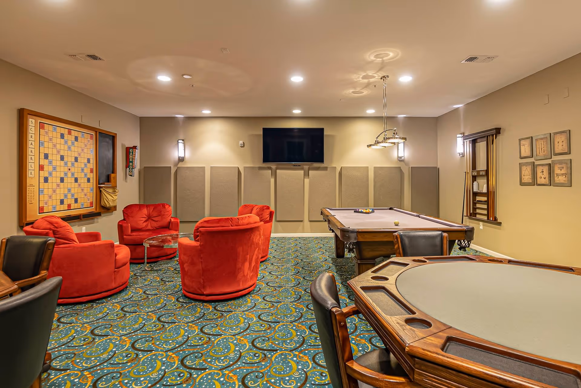 A game room with a pool table, a poker table, and four red cushioned chairs arranged around a small glass table. A large Scrabble board is mounted on the wall, and a flat-screen TV is mounted on the far wall above acoustic panels. The room has patterned carpet and warm lighting.