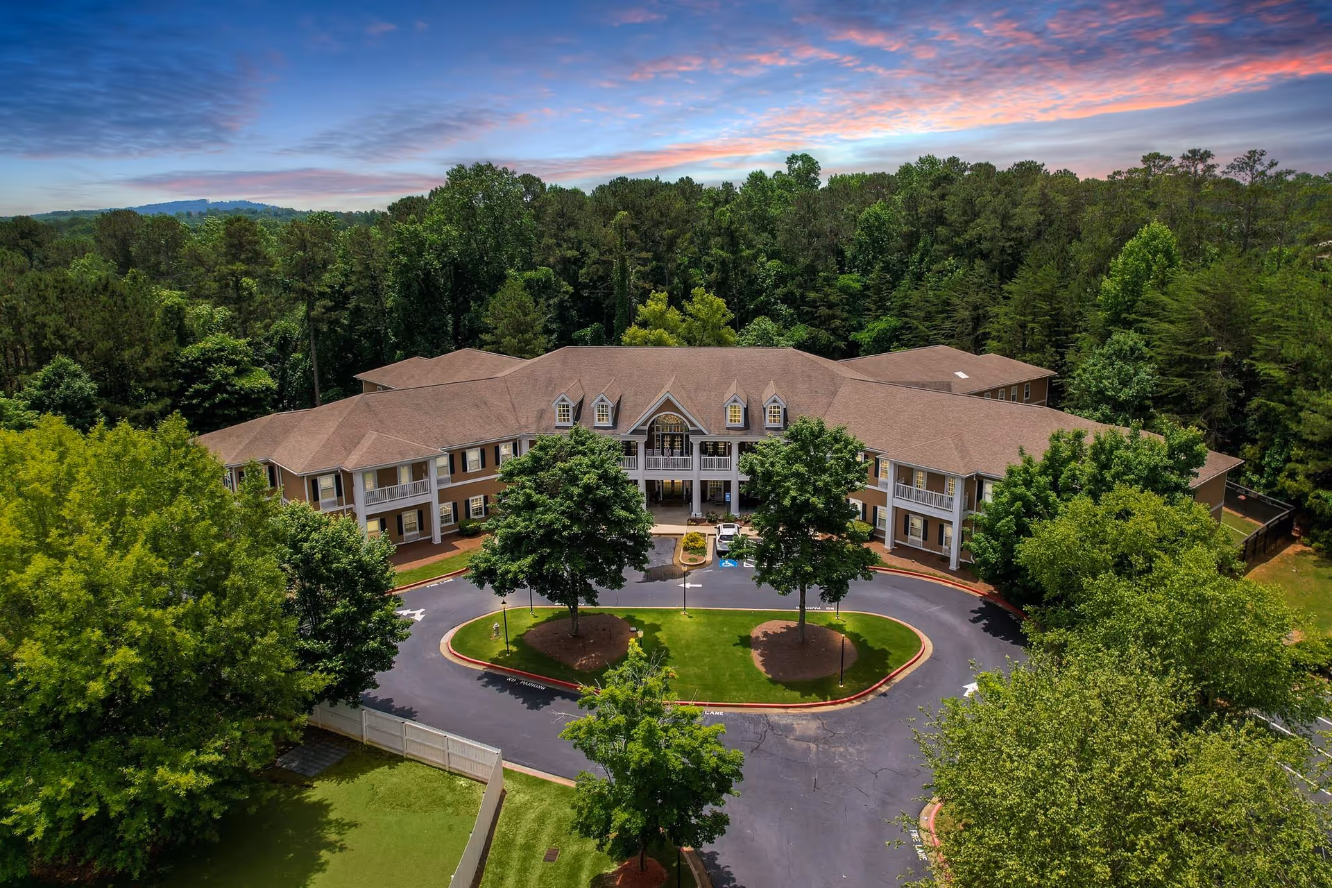 Aerial view of a large two-story senior living facility building surrounded by lush green trees and landscaping under a partly cloudy sky at sunset or sunrise. The building has a circular driveway with a small grassy island and several trees in front.