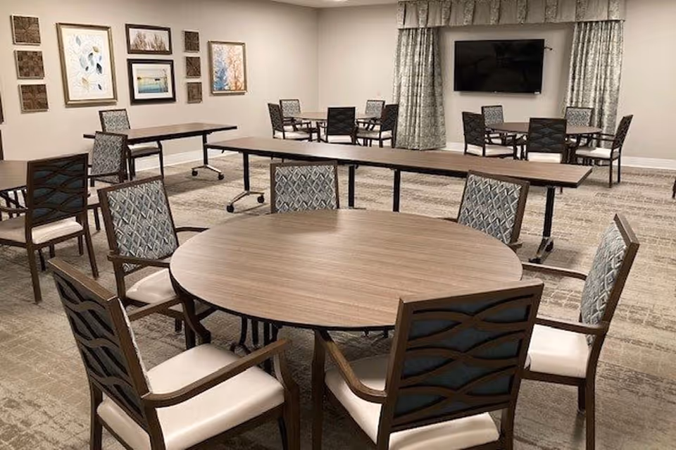A spacious room with multiple round and rectangular tables surrounded by cushioned chairs with patterned backs. The room has neutral-colored walls adorned with framed artwork and a large flat-screen TV mounted on the far wall with curtains on either side.