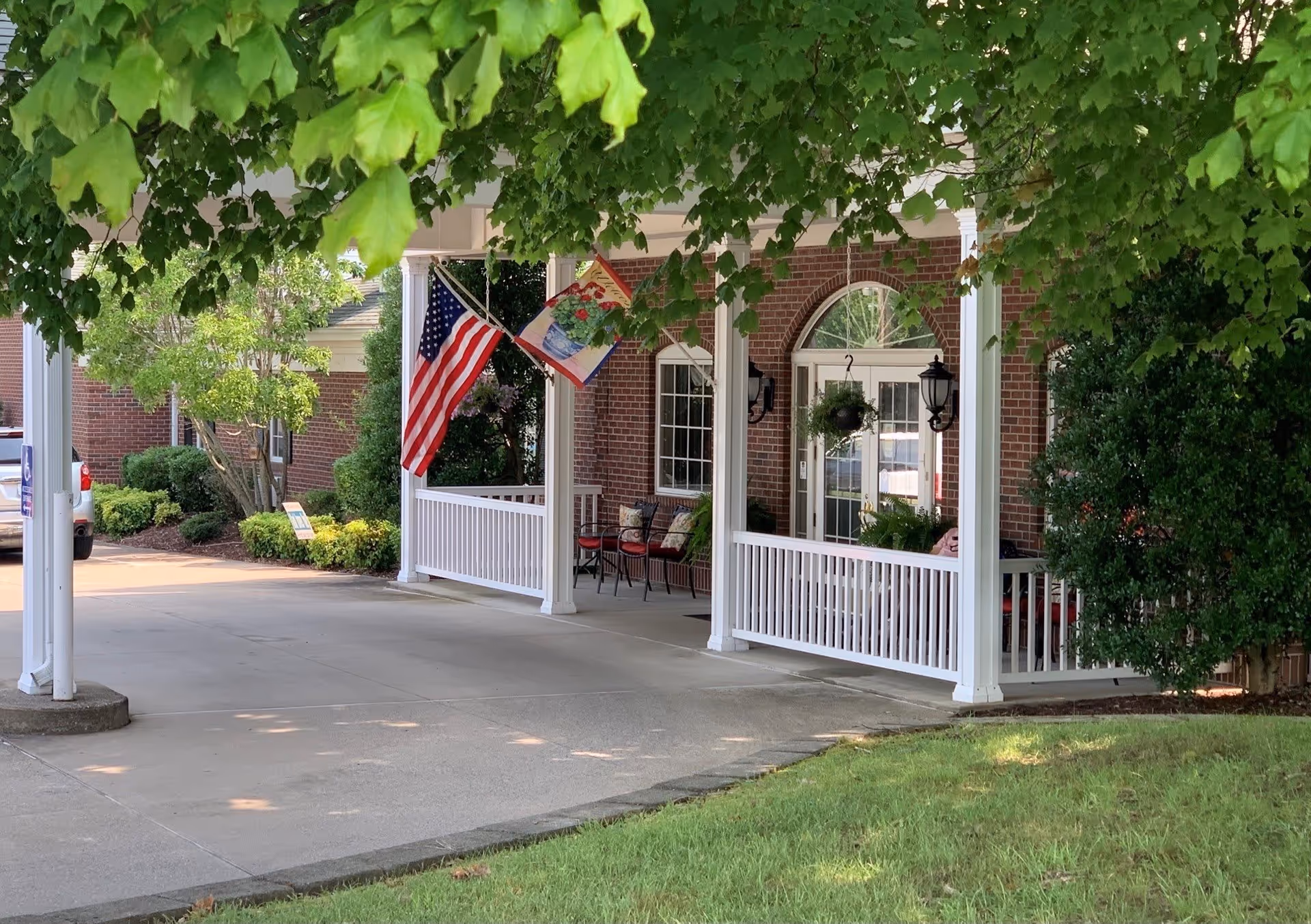 Covered driveway and porch at a brick senior living entrance with white columns, an American flag, chairs, and hanging plants.