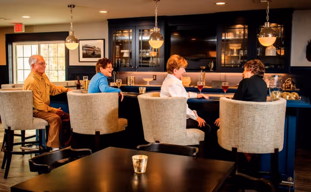 Four elderly people sitting on bar stools at a counter in a cozy, well-lit lounge area with pendant lights hanging above and glass cabinets in the background.