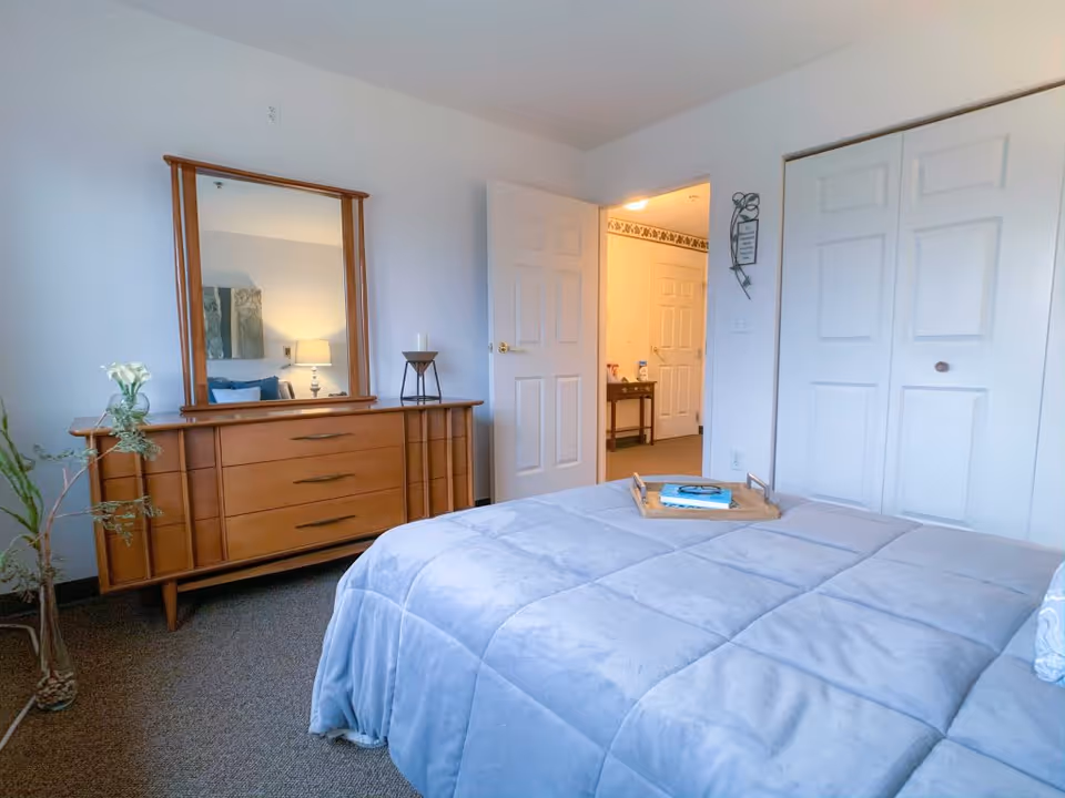 A bedroom with a light blue bedspread on the bed, a wooden dresser with a mirror, a small plant, and an open door leading to a hallway with another door and a small table with items on it.