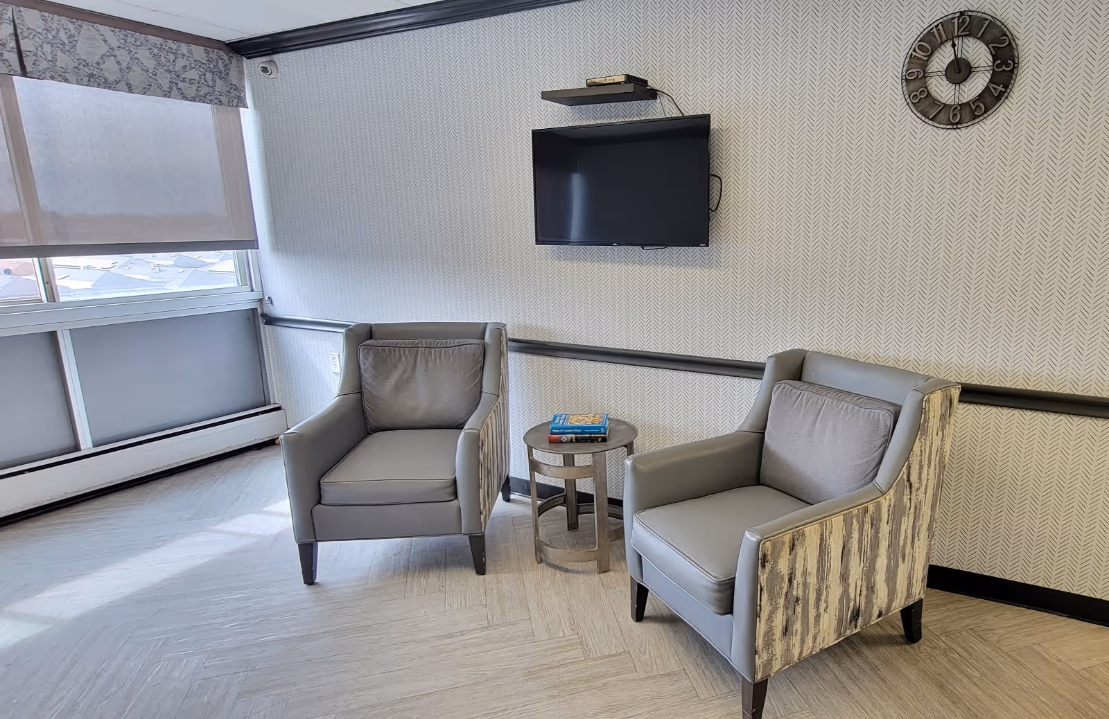 Two gray armchairs flank a small round table beneath a wall-mounted TV in a bright lounge area with large windows.