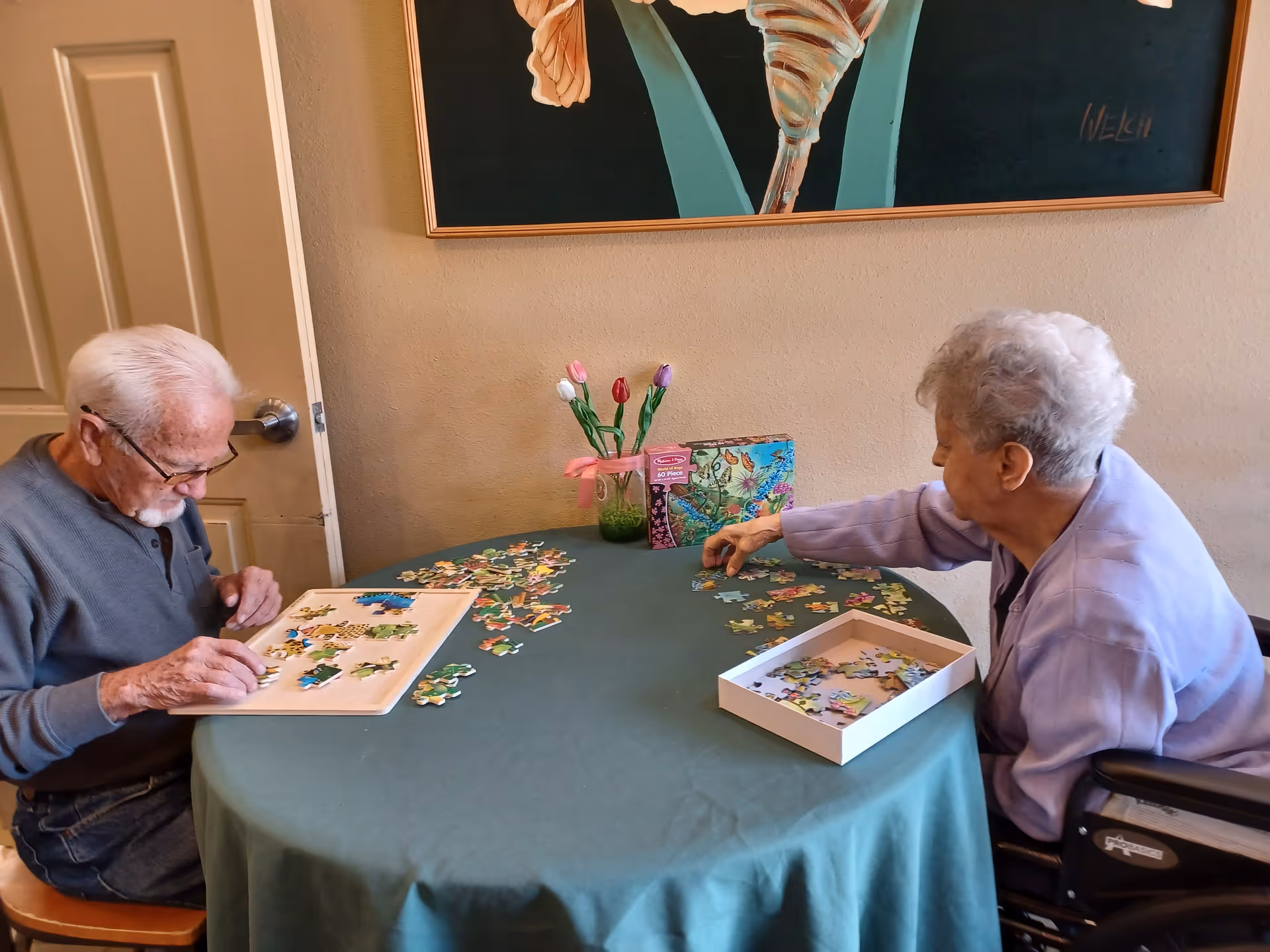 Two elderly individuals sitting at a round table covered with a green tablecloth, working on a jigsaw puzzle together. One person is seated on a chair while the other is in a wheelchair. There is a small vase with colorful artificial tulips and a puzzle box on the table. A large framed artwork is hanging on the wall behind them.