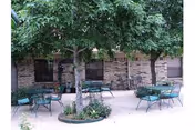 Outdoor patio area with several green metal tables and chairs arranged around two trees, adjacent to a brick building with windows.
