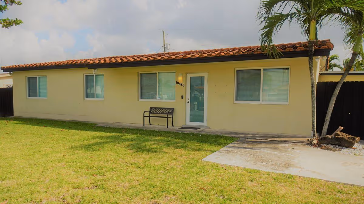 Single-story yellow building with a tiled roof, three front windows, a central glass door, a bench and a grassy lawn.