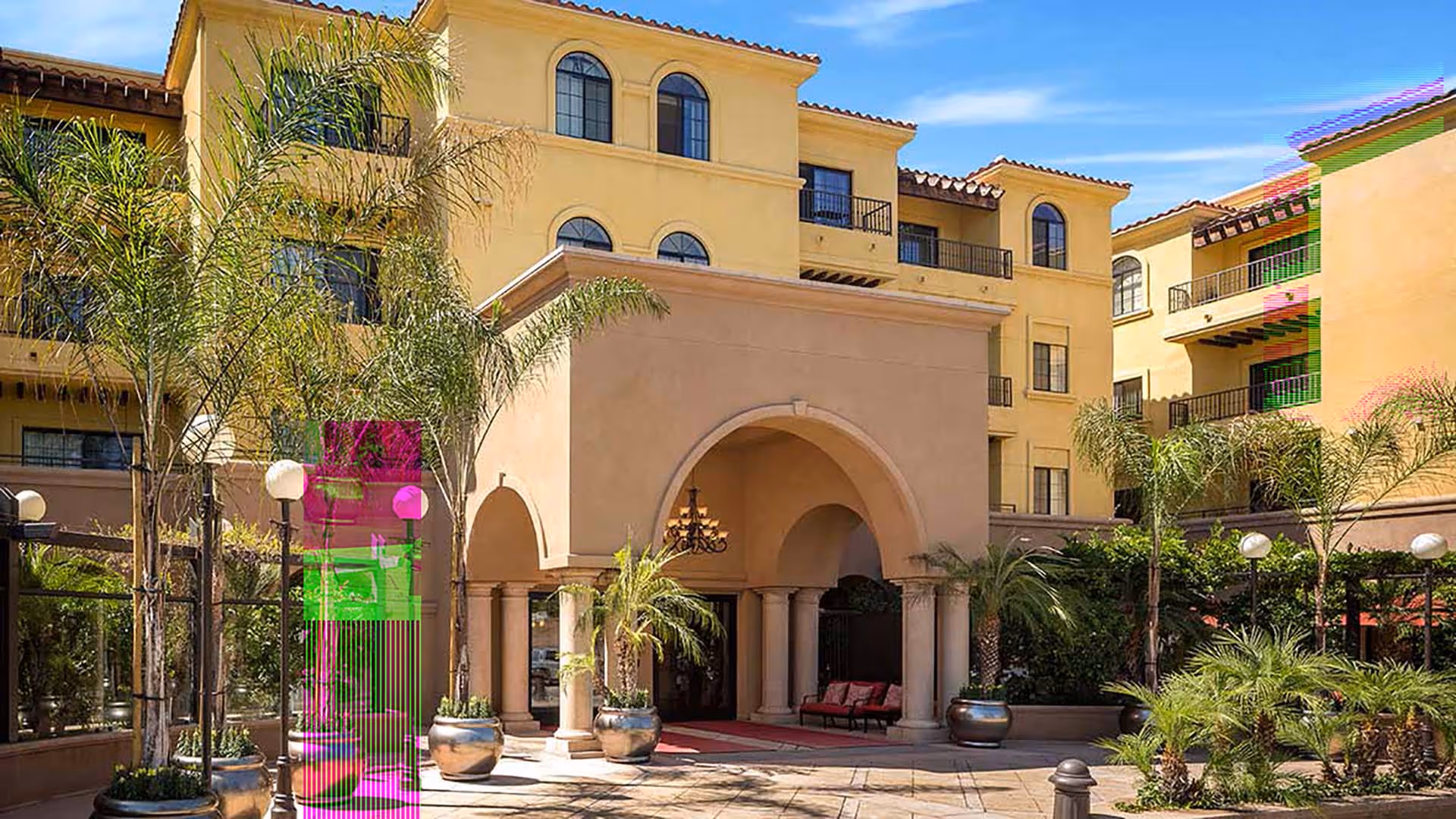Exterior view of Atria Tarzana, showing a Mediterranean-style building with arched entryway supported by columns, surrounded by palm trees and other greenery under a clear blue sky.