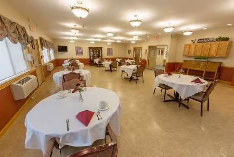 A bright dining room with multiple round tables covered in white tablecloths, place settings and chairs, and cabinetry along the walls.