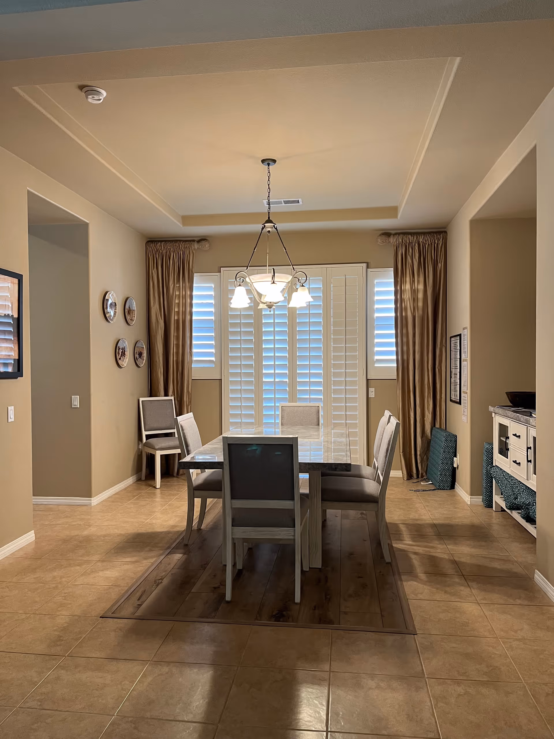 A dining room with a rectangular table surrounded by six chairs. The room has beige walls and tiled floors with a wooden area rug under the table. There are large windows with white plantation shutters and brown curtains. A chandelier with five lights hangs above the table. Decorative plates are mounted on the left wall, and a white sideboard with drawers and cabinets is on the right side of the room.