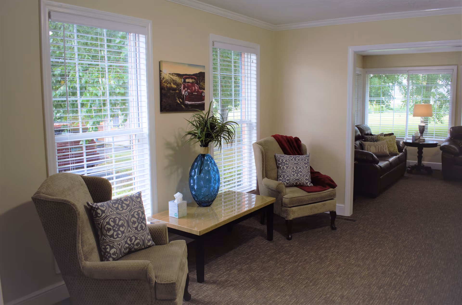 A cozy sitting area in an assisted living facility with two beige armchairs featuring patterned cushions and a red throw blanket. Between the chairs is a rectangular table with a large blue vase holding a green plant and a box of tissues. The room has large windows with white blinds letting in natural light. In the adjacent room, there are dark leather sofas, a side table with a lamp, and more windows showing greenery outside.