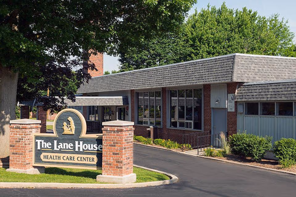 Exterior view of The Lane House Health Care Center building with a brick sign in front, surrounded by trees and bushes on a sunny day.