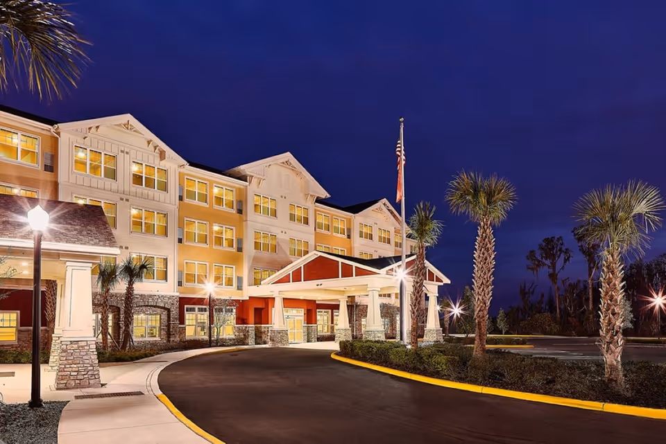Exterior view of Sonata Windermere senior living facility at night, showing a well-lit multi-story building with a covered entrance, palm trees, and a flagpole with an American flag.
