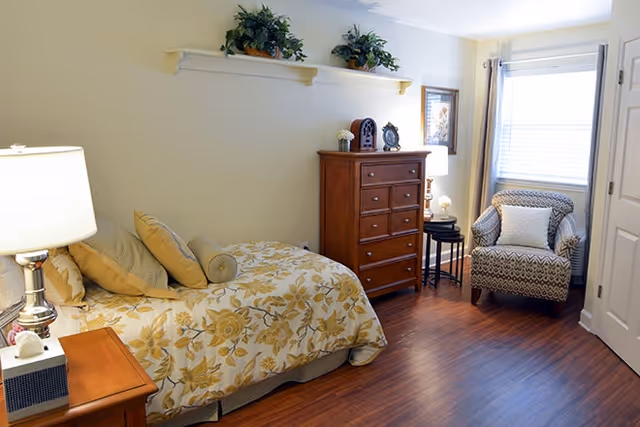A cozy bedroom with a single bed covered in a floral yellow and white bedspread, several pillows, a wooden nightstand with a lamp, a wooden chest of drawers, a patterned armchair with a white cushion, and a window with blinds and curtains. There are decorative plants on a wall shelf and framed artwork on the wall.