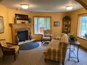 A cozy living room with a fireplace, a plaid sofa, two armchairs, a wooden chair, a small round rug, and a side table with plants. The walls are painted yellow and there are two windows letting in natural light.