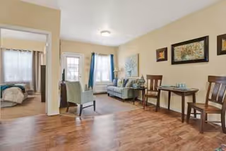 A cozy living area with wooden flooring featuring a small seating arrangement including a sofa, an armchair, and a small table with two chairs. The room is decorated with framed artwork on the walls and has windows with curtains allowing natural light to enter. A bedroom is visible through an open doorway.