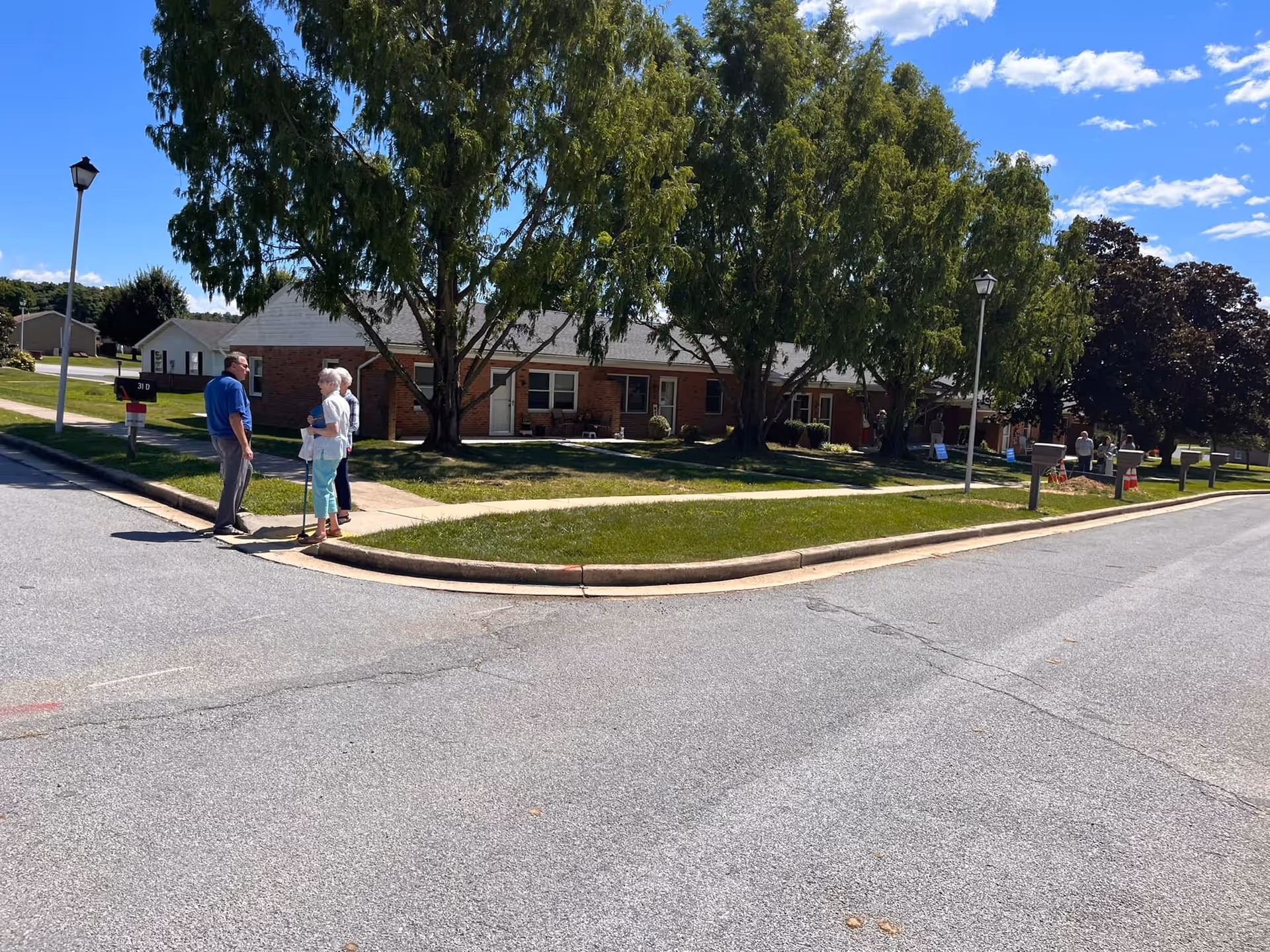 A sunny outdoor scene at Fahrney-Keedy Senior Living Community showing a paved road curving around a grassy area with large trees. Several people are standing and conversing near the sidewalk in front of a single-story brick building with multiple doors and windows. Mailboxes and street lamps line the sidewalk.