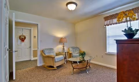 Small living room with two upholstered chairs, a glass coffee table, lamp, window with blinds and a dresser topped with a potted plant.