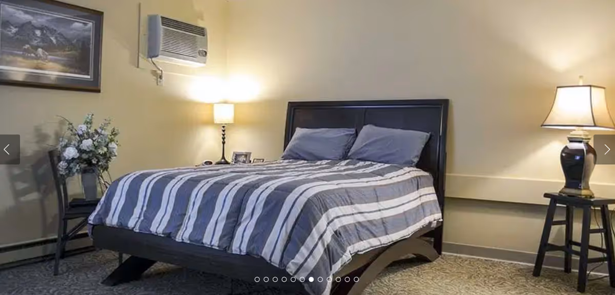 A bedroom with a dark wooden bed frame and a striped gray and white bedspread. There are two pillows on the bed. On the left side, there is a chair with a vase of white flowers and a framed picture on the wall above it. A wall-mounted air conditioner is above a lamp on a nightstand. On the right side, there is a small table with a lamp on it. The walls are painted a light beige color.
