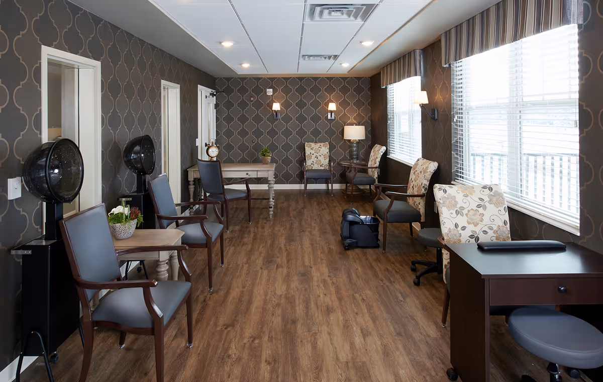 A well-lit room with wooden flooring and patterned dark wallpaper. The room contains several chairs with armrests, some with floral upholstery and others with plain dark cushions. There are two black hair dryer chairs on the left side, a small table with a plant, a desk with a lamp, and large windows with blinds on the right side. The ceiling has recessed lighting and air vents.