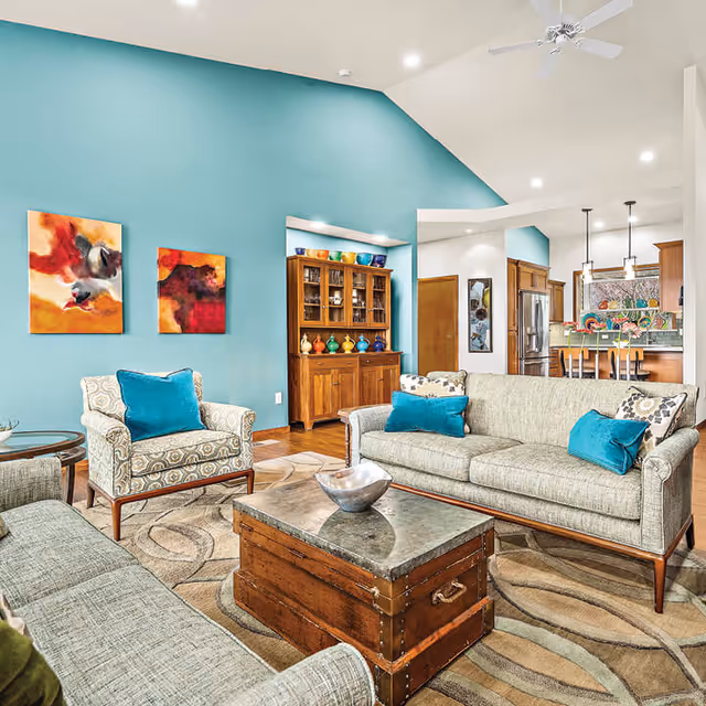 A bright and spacious living room with a high vaulted ceiling painted in teal. The room features two patterned armchairs and two sofas with teal accent pillows arranged around a wooden chest coffee table. A wooden cabinet with colorful pottery is against the wall, and the kitchen with a breakfast bar and pendant lights is visible in the background.