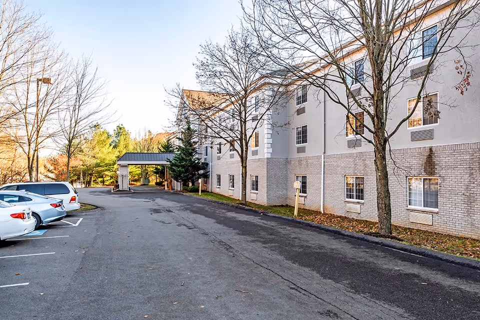 Exterior view of a senior living facility building with a parking lot on the left side. Several cars are parked in marked spaces, and leafless trees line the right side near the building. The building has multiple windows and a covered entrance area in the distance.