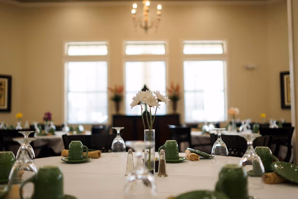 A dining room with tables set for a meal, featuring green cups and plates, upside-down wine glasses, rolled yellow napkins, salt and pepper shakers, and a vase with white flowers in the center. The room has large windows letting in natural light and a chandelier hanging from the ceiling.