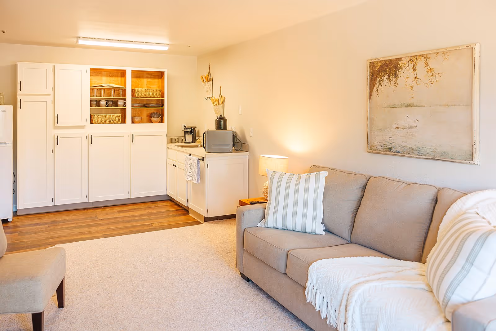 A cozy living area with a beige couch adorned with striped pillows and a white throw blanket. Next to the couch is a small wooden side table with a lamp. On the wall above the couch hangs a framed painting of two swans on a lake. The room features a small kitchenette with white cabinets, a microwave, a coffee maker, and some kitchen utensils. The floor transitions from carpet in the living area to wood in the kitchenette.