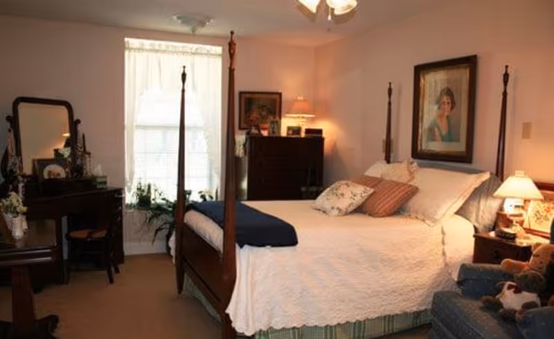 A cozy bedroom with a wooden four-poster bed covered in white bedding and multiple pillows. There is a wooden dresser with a lamp and framed pictures, a vanity with a mirror and chair, a window with sheer curtains, and a comfortable armchair with stuffed animals. The walls are decorated with framed artwork.