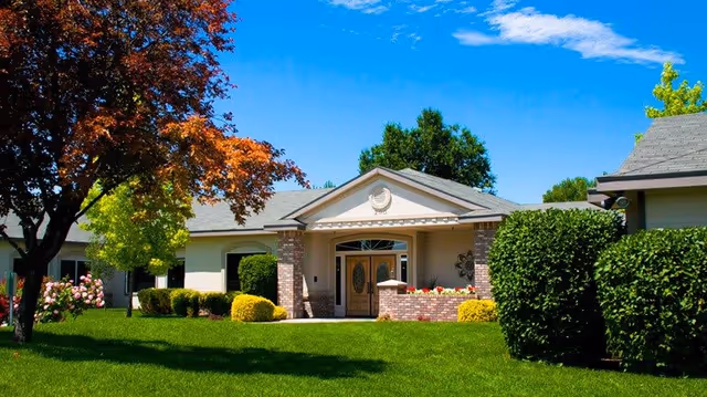 Front exterior view of a single-story assisted living facility building with a well-maintained green lawn, colorful trees, and shrubs under a bright blue sky with a few clouds.