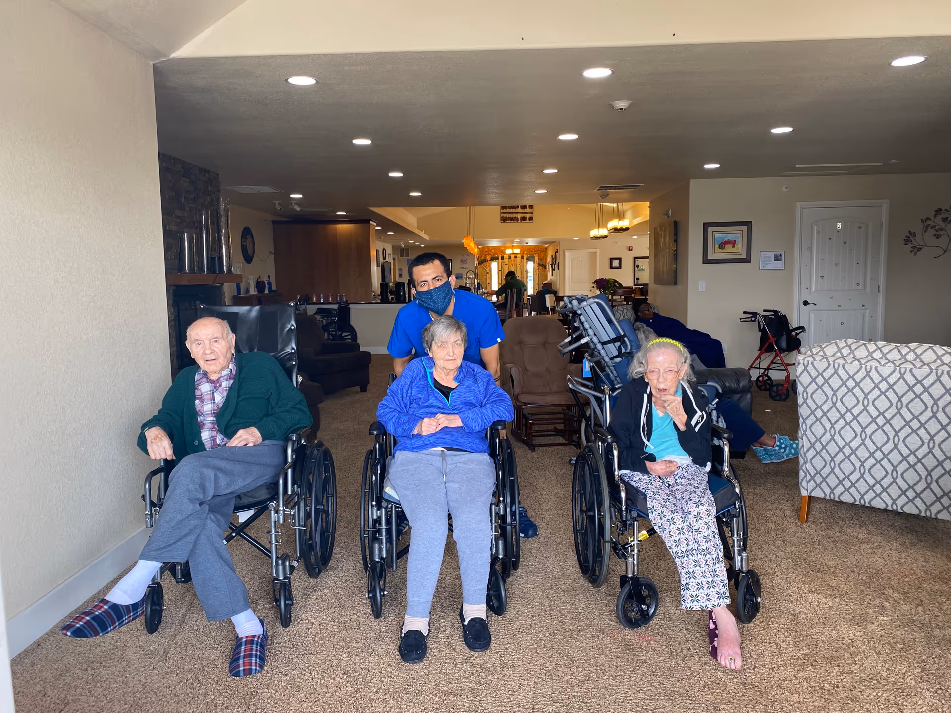 Three residents in wheelchairs with a staff member in a communal living room of an assisted living facility.