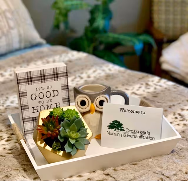 A white tray on a bed with a plaid blanket, holding a small potted succulent plant, an owl-shaped mug, a decorative sign that reads 'It's so good to be home,' and a welcome card from Crossroads Nursing & Rehabilitation.