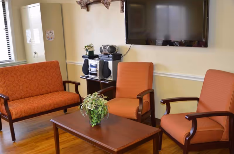 Small sitting area with orange upholstered chairs and a loveseat around a wooden coffee table with a potted plant, a stereo cabinet, and a wall-mounted TV.