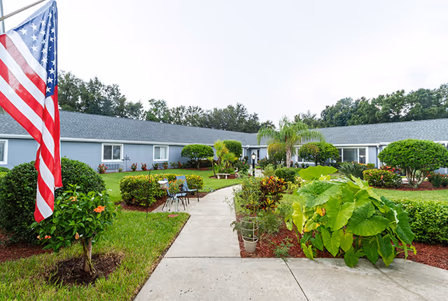 Well-maintained courtyard with a paved walkway, chairs, landscaped plants and an American flag in front of a single-story assisted living building.