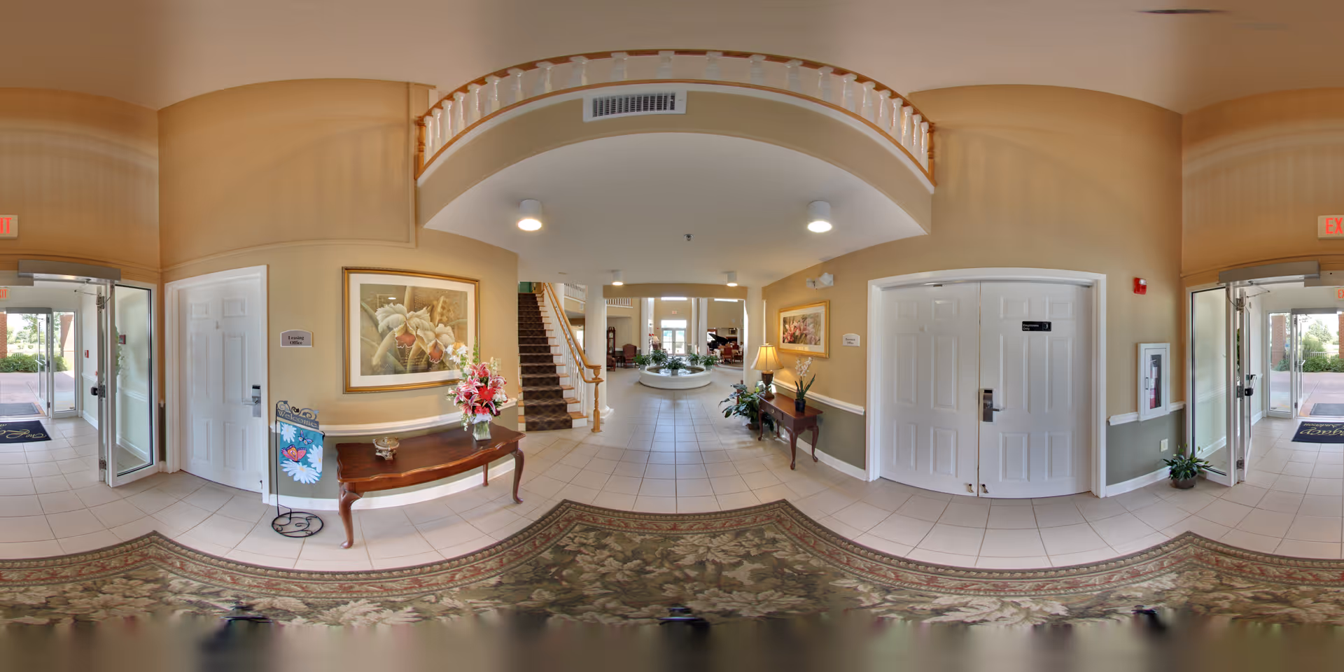 A spacious and well-lit senior living facility lobby with beige walls and tiled floors. There are two wooden tables with flower arrangements and framed artwork on the walls. A staircase with a wooden handrail leads to an upper floor. Double doors and glass entrance doors are visible, along with a patterned area rug on the floor.