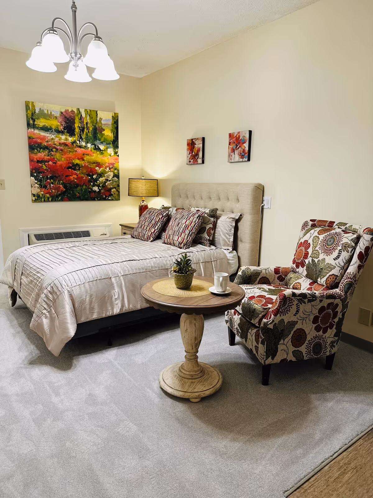 A cozy senior living bedroom featuring a neatly made bed with multiple patterned pillows, a floral upholstered armchair, and a round wooden side table with a small plant and a cup on a saucer. The room has light-colored walls adorned with colorful paintings, a bedside lamp, and a ceiling light fixture.