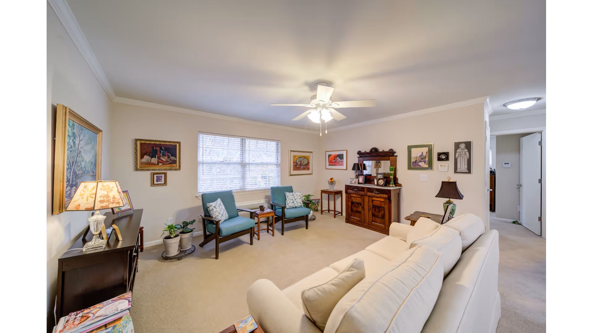 Bright, traditionally furnished living room with a beige sofa, two armchairs, wooden cabinets, artwork on the walls, and a ceiling fan.