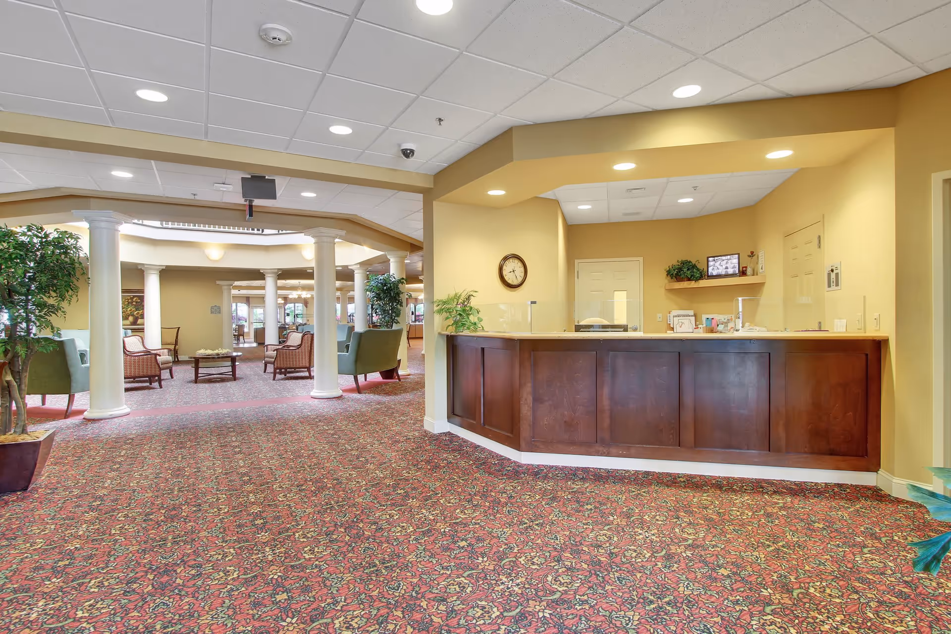 Interior view of a senior living facility lobby area featuring a reception desk with a clock on the wall behind it, potted plants, and a spacious seating area with armchairs and small tables. The floor is covered with a patterned carpet, and the ceiling has recessed lighting.