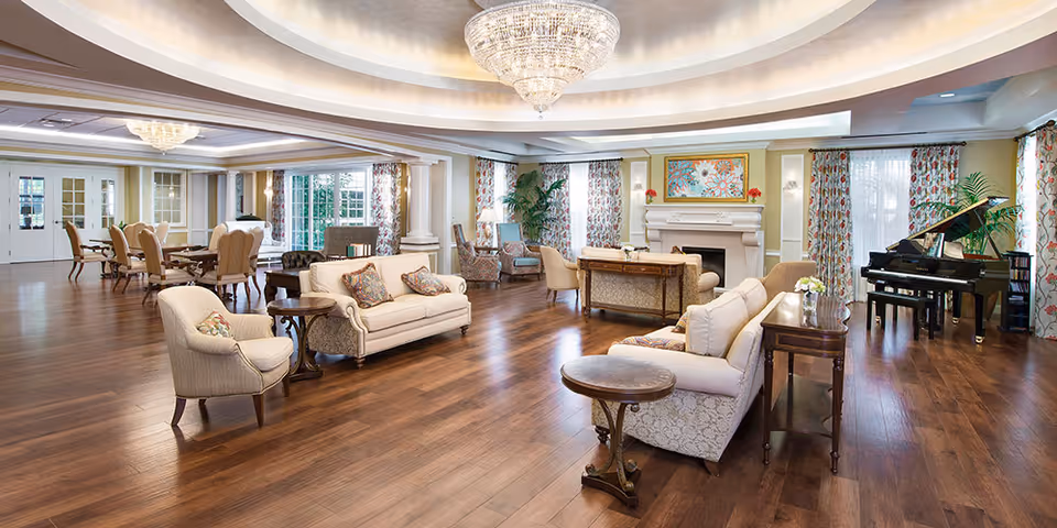 Spacious and elegant senior living common area with hardwood floors, multiple seating arrangements including sofas and armchairs, a grand piano near large windows with floral curtains, a fireplace with artwork above it, and a chandelier hanging from the ceiling.