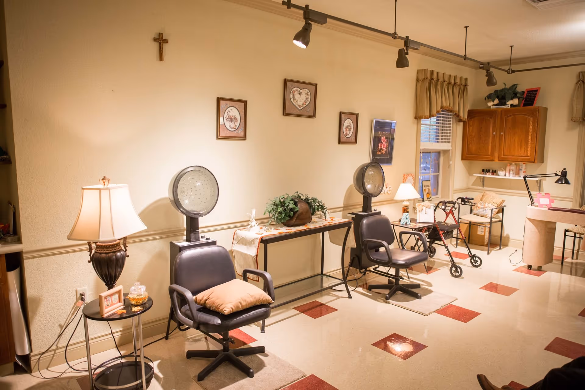 Interior of a senior living facility room with two black salon chairs under hair dryers, a small table with a lamp and framed photo, a walker, a chair with a cushion, wall decorations including framed pictures and a cross, and a cabinet with various items on top and shelves.