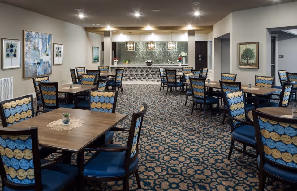A spacious dining room with multiple wooden tables and chairs featuring blue and yellow patterned upholstery. The room has a patterned carpet and light-colored walls adorned with framed artwork. At the far end, there is a counter with a textured backsplash and pendant lights hanging above it.