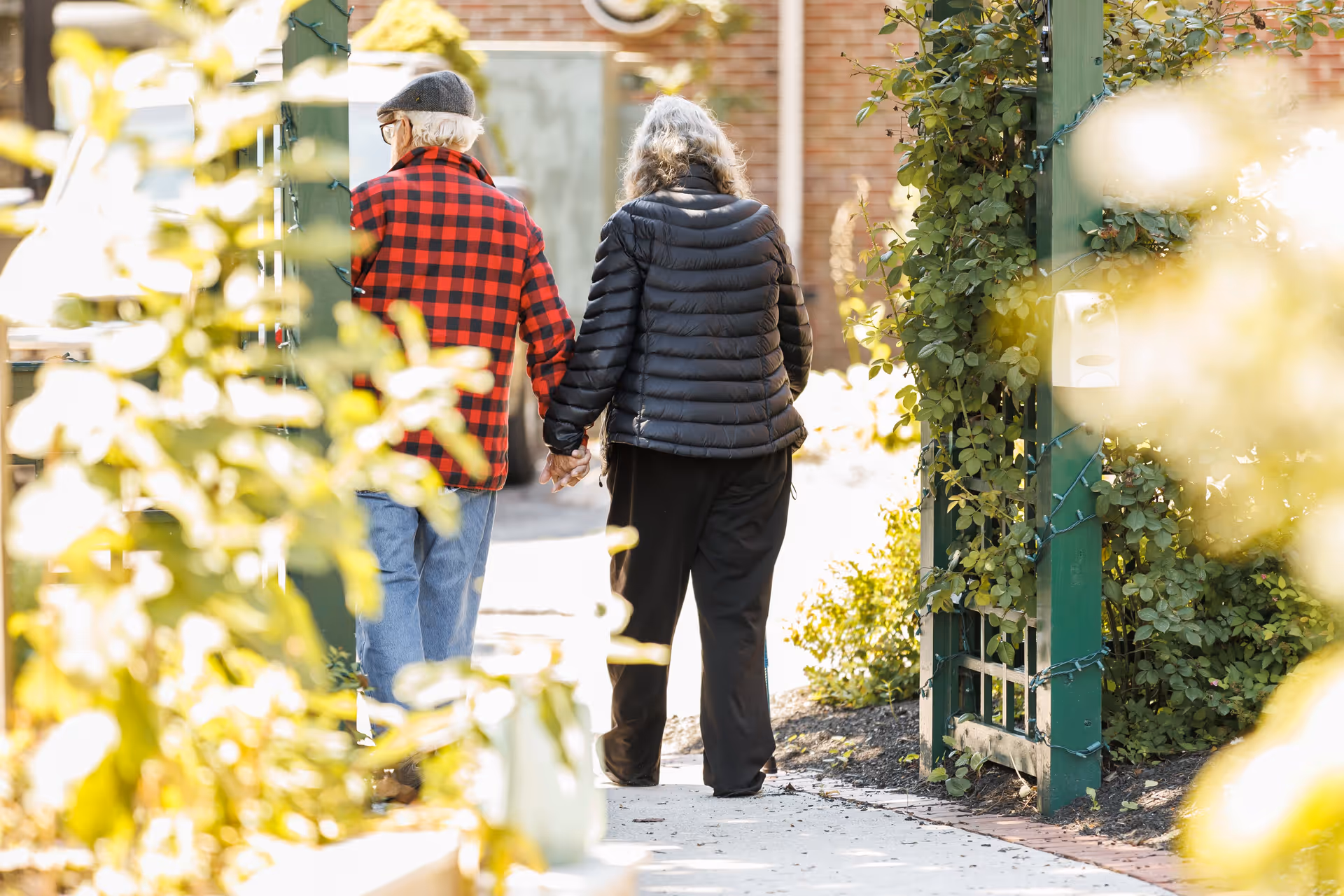 An elderly couple holding hands and walking away through a garden pathway with greenery and sunlight around them.