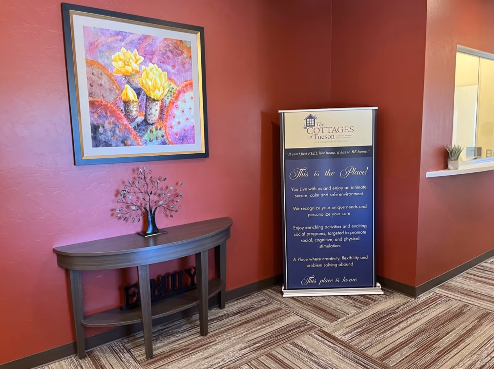 Interior reception area with red walls, a framed cactus painting, a small console table and a standing sign for The Cottages of Tucson.