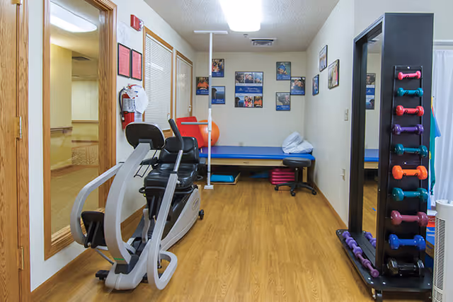 A small exercise room with wooden flooring featuring a recumbent exercise bike, a rack of colorful dumbbells, an exercise mat on a bench, and various exercise balls. The walls are decorated with framed pictures and certificates, and there is a large mirror on one side.