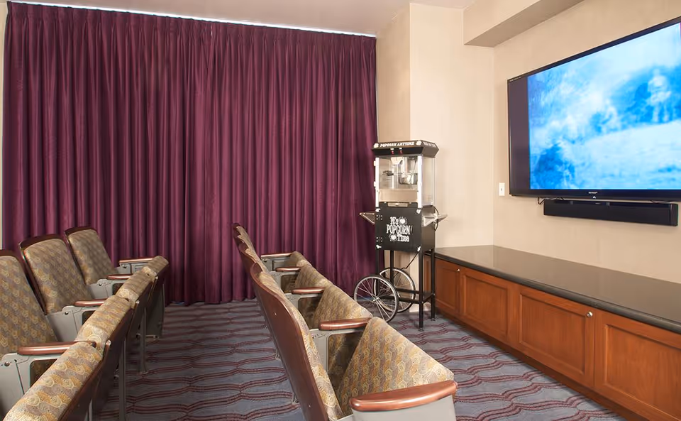 A small theater room with two rows of patterned cushioned chairs facing a large flat-screen TV mounted on the wall. There is a popcorn machine on a stand next to the TV, and the room has purple curtains covering the back wall and a carpeted floor with a wavy pattern.