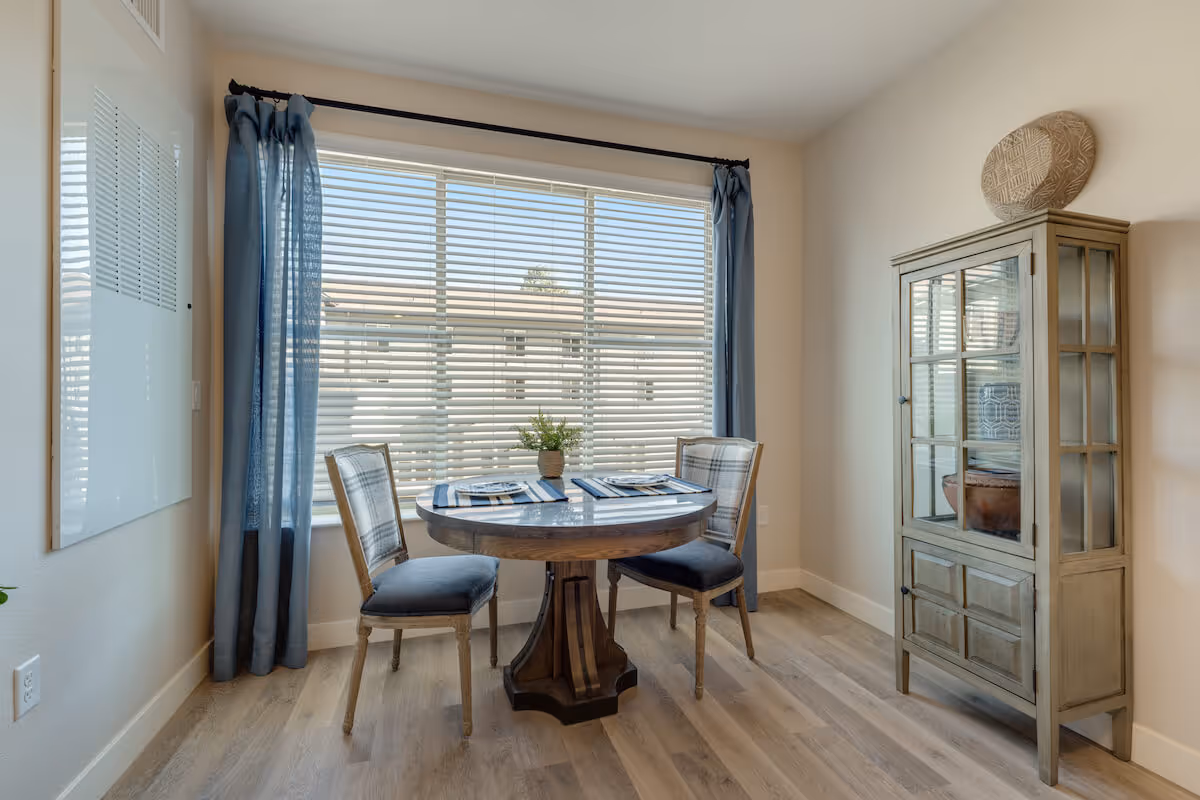Bright dining nook with a round wooden table set for two, two upholstered chairs, a glass-front cabinet, and a large window with blue curtains.