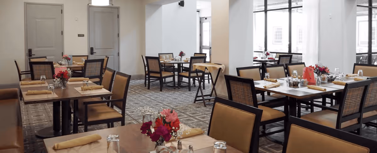 A dining room with several tables set with napkins, glasses, and small flower arrangements. The room has large windows letting in natural light and multiple chairs around each table.