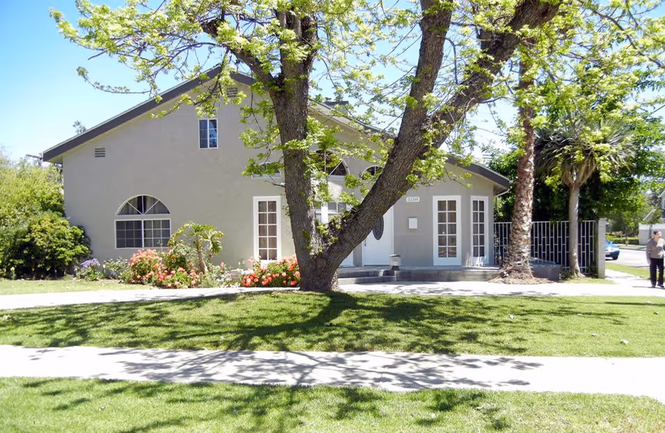 Exterior view of a single-story building with light gray walls and white-framed windows and doors. There is a large tree with green leaves in front of the building, along with a well-maintained lawn and flower beds with colorful flowers. A sidewalk runs in front of the lawn, and a person is visible walking on the right side near the street.