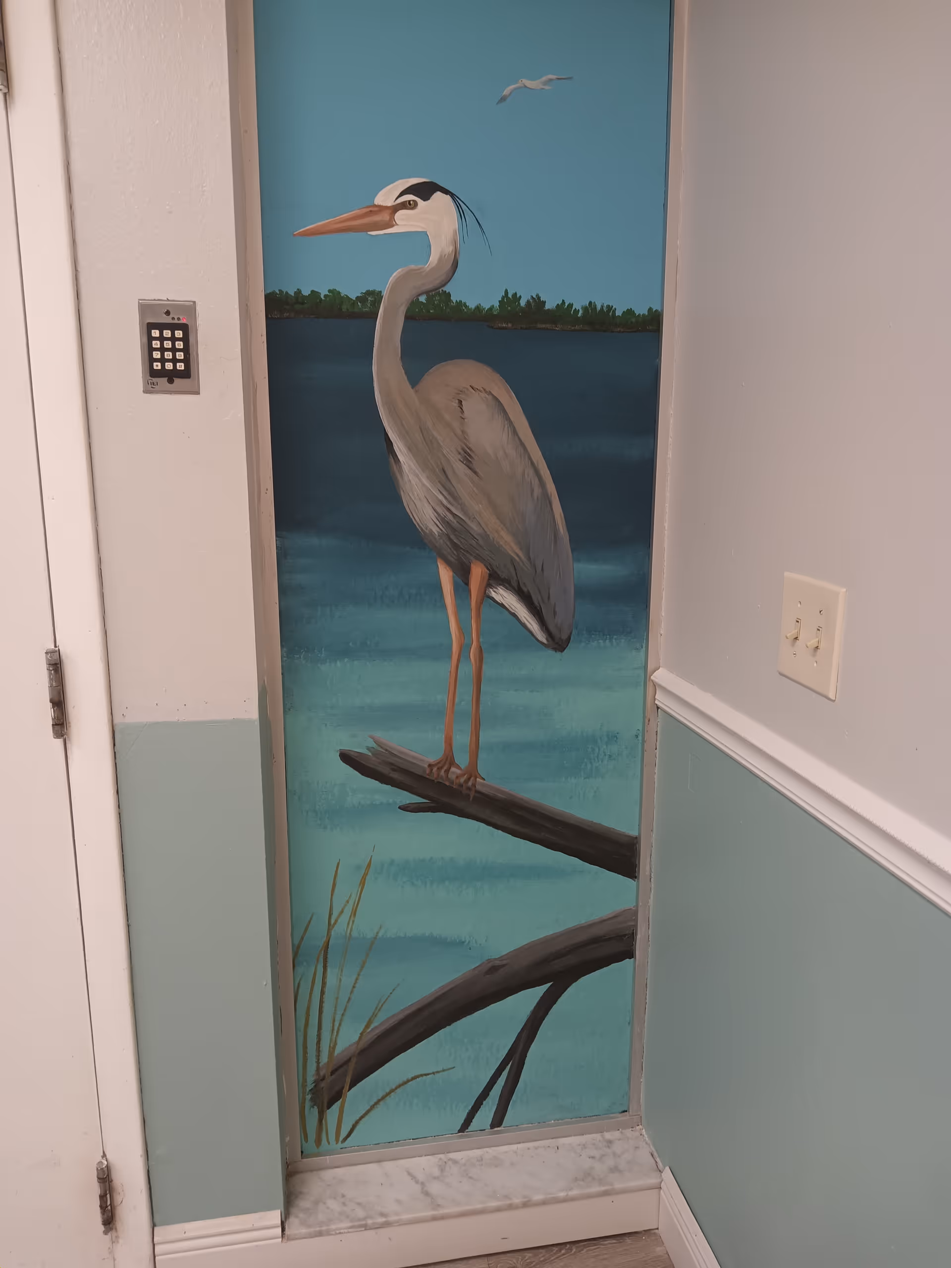 A painted mural of a heron standing on a branch over water with a background of trees and a flying bird, located on a narrow wall section between a door and a light switch in an indoor hallway.